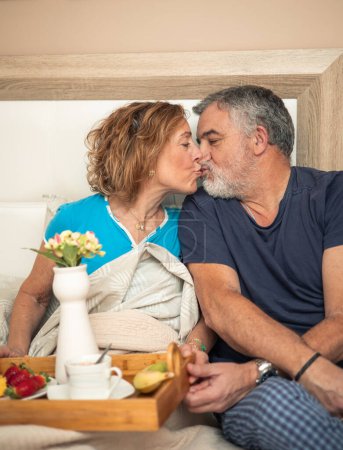 Dieser zärtliche Moment fängt ein älteres Paar in einer liebevollen Umarmung ein, die beim Frühstück im Bett einen Kuss teilt. Ihre liebevolle Geste spricht Bände über die tiefe Verbundenheit, die sie auch nach Jahren der Kameradschaft teilen. 