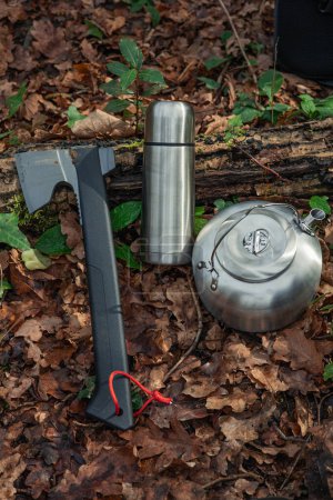 In the heart of the forest, a set of essential tools rests on the ground: an axe, a metal thermos, and a camping kettle. These items symbolize preparation and connection with nature, ready to accompany adventurers on their expeditions and provide com