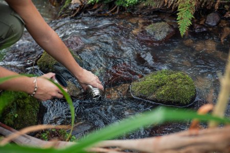 In the serene heart of the forest, skilled hands dip into a crystal-clear river, gathering water in a metal container. This image captures the essence of human connection with nature, emphasizing the importance of autonomy and sustainability in our j