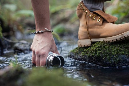 Gros plan de la main et du pied avec récipient métallique recueillant l'eau d'une rivière au milieu d'une forêt.