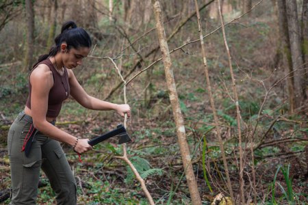"In this compelling image, a fearless adventurer undertakes the task of constructing a shelter in the heart of the forest, using bushcraft and survival skills to chop branches from a tree with an axe. Her determination and expertise underscore the im