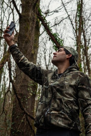 man in camouflage, gazing up with cellphone in hand, endeavors to find signal amidst towering forest canopies.
