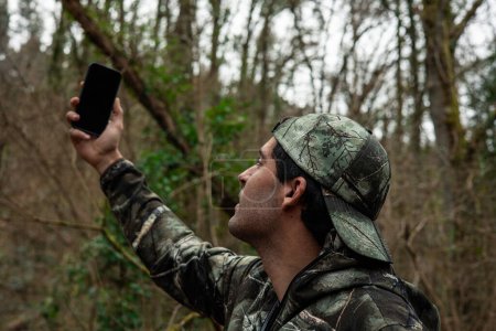 A man in camouflage struggles to find cell signal, surrounded by dense forest, with cellphone raised in hand