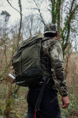 Vue arrière d'un homme debout dans la forêt dense, portant un sac à dos contenant un récipient en métal d'eau et une hache. Il semble vigilant au milieu de la végétation luxuriante