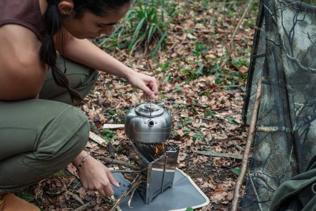 Image of a young adventurer cooking in the forest, holding a metal kettle over a portable bushcraft stove, demonstrating outdoor cooking skills and wilderness survival