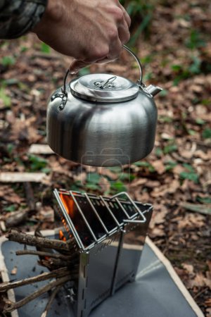 vertical portrait Closeup shot of a hand placing a metal kettle on a survival stove in the middle of the forest, demonstrating outdoor survival skills and preparing water in the wilderness