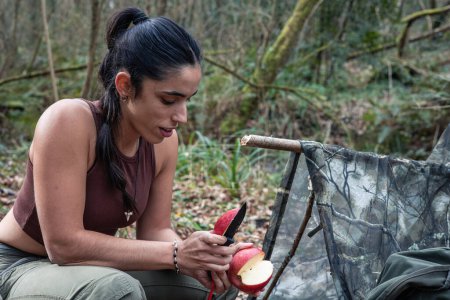 Photograph of a young explorer girl seen enjoying a freshly cut apple by her shelter in the middle of the forest. The image captures a moment of serenity and connection with nature as she savors her snack and relaxes in her natural surroundings.