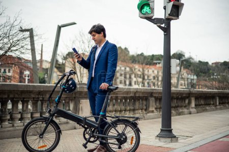 A modern urban commuter, dressed in business attire, efficiently multitasks by sending messages on his smartphone while holding his electric folding bike and helmet. This image exemplifies the seamless integration of technology and sustainable transp