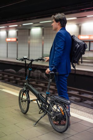 Ein stylischer junger Geschäftsmann wartet mit seinem Rucksack und einem faltbaren Elektrofahrrad an der U-Bahn-Station und demonstriert sein Engagement für einen umweltfreundlichen Lebensstil und nachhaltigen Verkehr in der Stadt. Das Bild spiegelt die Kombination von aktivem Mobbing wider