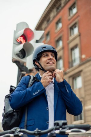 Ein Geschäftsmann im Anzug wird dargestellt, wie er seinen Helm für eine sichere Fahrt mit seinem Elektrofahrrad in der Stadt anschnallt. Dieses Bild unterstreicht die Bedeutung von Sicherheit und umweltbewussten Transportmöglichkeiten in städtischen Umgebungen