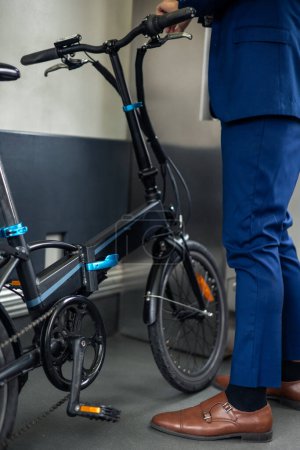 A closeup shot captures the electric folding bike and the legs of a businessman in a suit inside the metro. This image highlights the integration of sustainable transportation solutions into urban commuting, emphasizing the businessman's eco-friendly
