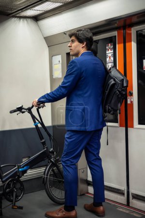 Ein Geschäftsmann im Anzug ist zu sehen, wie er mit einem Rucksack und einem elektrischen Faltrad für einen nachhaltigen Stadtverkehr zur Arbeit in der U-Bahn pendelt. Dieses Bild zeigt die Integration umweltfreundlicher Mobilitätslösungen in die 