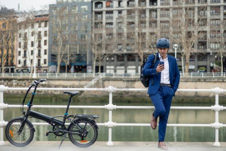 A young businessman, dressed in a suit and carrying a backpack, leans against the railing by the river with his electric bike beside him. He is engaged in texting on his smartphone, possibly contemplating his day or communicating with colleagues whil