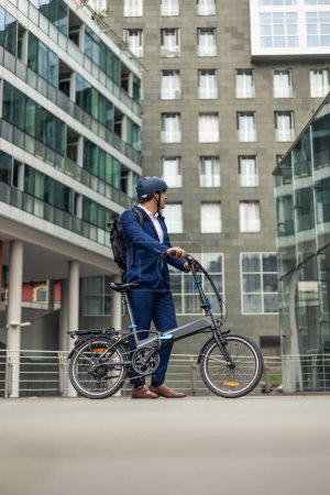 Ein Geschäftsmann im Anzug steht mit seinem Elektrofahrrad inmitten von Bürogebäuden im städtischen Geschäftsviertel und betont die Bedeutung nachhaltigen Verkehrs in der Stadt