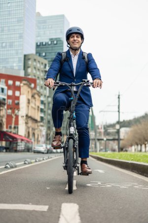 retrato vertical vista frontal de un hombre de negocios sonriente en traje pedaleando su bicicleta eléctrica plegable a lo largo de un carril dedicado a la bicicleta en la ciudad, promoviendo el transporte urbano sostenible y de seguridad