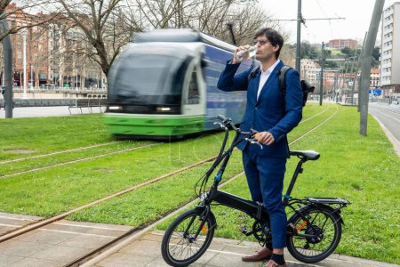 suited businessman takes a momentary pause from his bustling commute, sipping water from a glass bottle while standing beside his folded electric bike. With sunglasses on, he embraces the eco-friendly lifestyle amidst the city's vibrant streets, as a