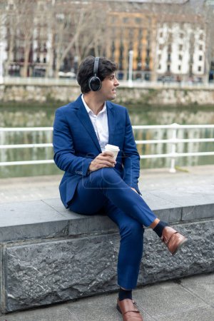 Ein männlicher Geschäftsmann in blauem Anzug und Kopfhörer sitzt am Fluss in der Stadt und genießt einen Kaffee zum Mitnehmen.