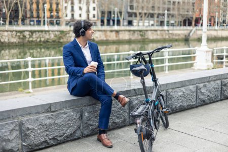 copyspace A businessman in a blue suit and headphones takes a moment to relax by the river in the city, next to his folding electric bike, while sipping on a takeaway coffee