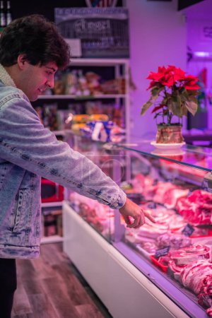 Photo for Vertical portrait A closeup shot captures a customer in a butcher shop, carefully pointing at the assortment of meats showcased on the counter. With a focused gaze, they make their selections from the array of quality cuts and specialties available - Royalty Free Image
