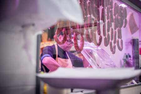 Ein Metzger mit einer Vakuumsiegelmaschine, während im Vordergrund Würstchen und Chorizos von der Decke der Metzgerei hängen. Die Szene unterstreicht die traditionellen Konservierungsmethoden und die Handwerkskunst des Metzgers