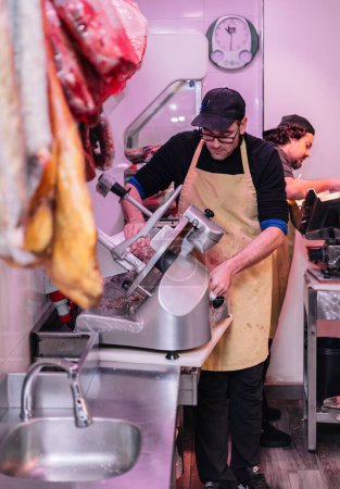 Photo for Vertical portrait two butchers wearing yellow aprons using their electric meat slicers in their butcher shop. The scene reflects the efficiency and professionalism of the butchers in their workflow, ensuring precise and quality cuts for their custome - Royalty Free Image