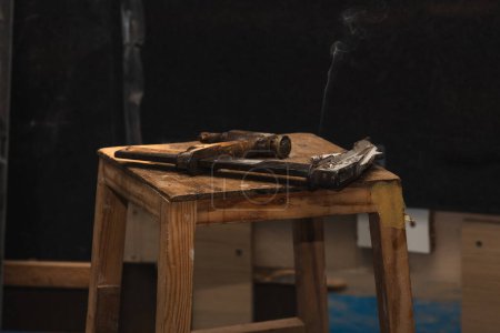 abrazadera de carpintero vintage de primer plano descansando junto a un cigarrillo humeante en un banco de trabajo de madera rústica en un taller de carpintería tradicional. La escena emana nostalgia y artesanía, capturando la esencia de una embarcación atemporal en medio del humo arremolinado