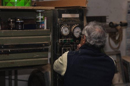 elderly carpenter, seen from behind, as he observes the gauges of an old wood press machine to monitor time and pressure in his workshop. The scene embodies a blend of experience and precision, highlighting the timeless craftsmanship and attention to