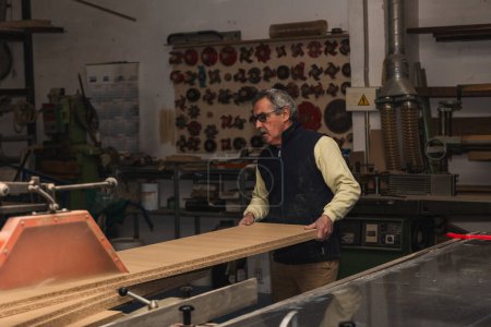 Foto de Copyspace anciano carpintero maniobras de una sierra circular en su taller de carpintería en esta imagen. El artesano sostiene firmemente una gran tabla de madera mientras la empuja a través de la mesa de cortar, mostrando su habilidad y experiencia en el comercio. En el fondo. - Imagen libre de derechos