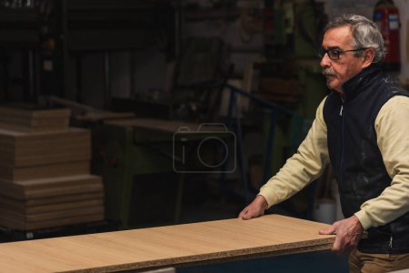 Auf diesem Bild greift ein älterer Tischler in seiner Tischlerei nach einer großen Holzplatte. Mit konzentriertem Gesichtsausdruck und Brille demonstriert der erfahrene Handwerker sein Können und sein Fachwissen in der Holzbearbeitung. Die Szene verströmt ein Gefühl von Tristesse