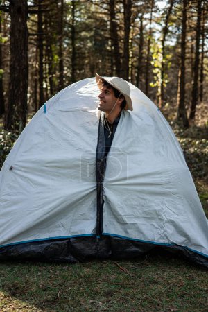 Ein lächelnder Camper mit Forscherhut guckt an einem Sommermorgen aus dem Zelt und umarmt die Ruhe der Natur mit fröhlichem Auftreten.
