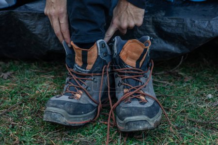 Closeup shot of a man putting on a pair of trekking boots covered in dirt while standing on grass, getting ready for an outdoor adventure