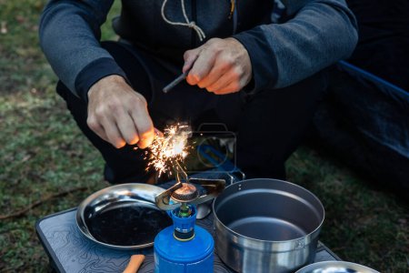 Ein Camper ist zu sehen, wie er versucht, einen Campingkocher auf dem Campingplatz in der Natur anzuzünden, wobei Funken vom Feuerstein fliegen, während er sich inmitten der Wildnis auf das Abendessen vorbereitet.
