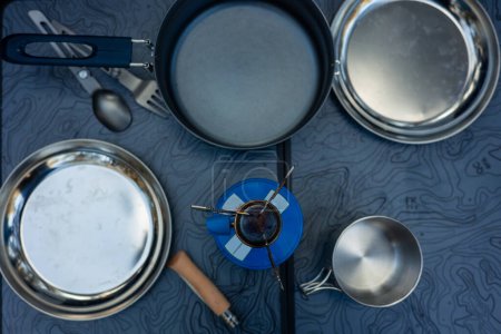 Eine Overhead-Perspektive zeigt einen Campingtisch mit verschiedenen Kochutensilien für die Zubereitung von Mahlzeiten auf dem Campingplatz, darunter einen Campingkocher, Teller, Töpfe, Besteck und eine Metalltasse.
