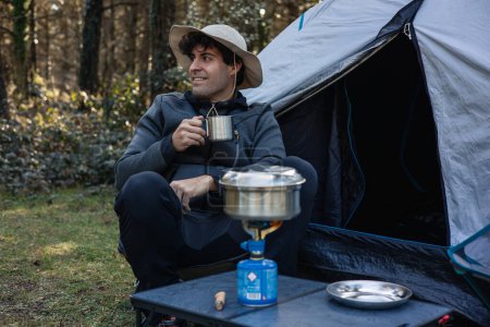 Vertikales Porträt Ein Mann beim Zelten sitzt vor seinem Zelt und trinkt Kaffee aus seiner Metalltasse, während er inmitten der Natur sein Essen auf dem Campingkocher zubereitet
