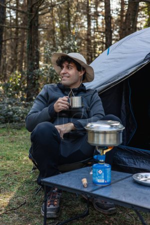 vertical portrait solitary camper sits by his tent, enjoying a moment of tranquility while sipping coffee, surrounded by the sights and sounds of the natural world, a testament to the beauty of outdoor exploration and wildlife encounters