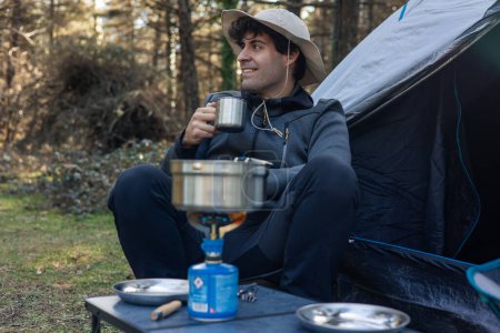 Man sieht einen Mann in der Wildnis campieren, vor seinem Zelt sitzen und Kaffee schlürfen, während sein Essen auf dem Campingkocher kocht.