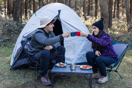 Multikulturelles Paar aus dem vorherigen Bild lächelt jetzt und prostet freudig mit seinen Tassen auf dem Campingplatz zu und teilt einen Moment des Glücks und der Verbundenheit während ihres Outdoor-Abenteuers