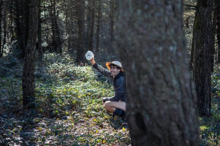 young camper wearing an explorer hat is shown defecating behind a tree trunk in the middle of the forest, while holding a roll of toilet paper in his hand