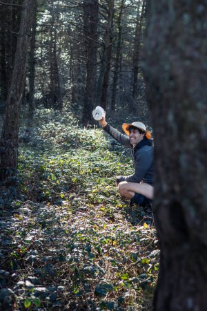 Ein junger Camper demonstriert Hygienepraktiken, indem er sich im Wald hinter einem Baumstamm entledigt und dabei eine Rolle Toilettenpapier in der Hand hält. Diese Geste unterstreicht die Wichtigkeit, die natürliche Umwelt und Umwelt zu respektieren.