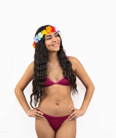 Fröhliche junge Latina posiert selbstbewusst in einem Bikini mit den Händen auf den Hüften und einer Blumenkrone, strahlt Freude aus und genießt die sommerliche Stimmung auf weißem Hintergrund