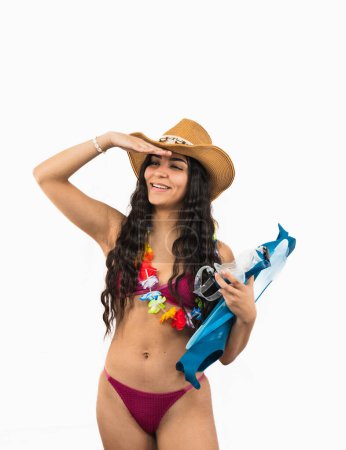 junge Latino-Frau im Bikini hält Tauchflossen, während sie ihre Augen vor der Sommersonne mit der Hand vor weißem Hintergrund abschirmt