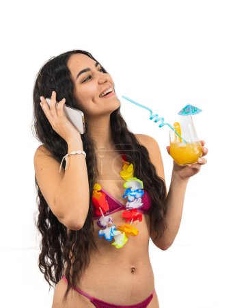 young Latin woman wearing a bikini is talking on her smartphone while holding a summer cocktail in her other hand. She appears relaxed and engaged, enjoying a sunny beach day on white background