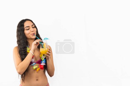 Téléchargez les photos : Copyspace brunette femme latine portant un bikini et collier de fleurs profite d'un cocktail rafraîchissant lors d'une chaude journée de plage d'été, rayonnant de détente et de plaisir sur fond blanc - en image libre de droit