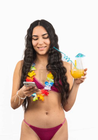 young Latina woman wearing a bikini is seen texting on her smartphone while holding a summer orange soda in her other hand. She appears relaxed and engaged on white background