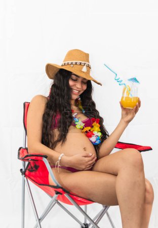 regnant Latin girl wearing a bikini is seen sitting on a beach chair, enjoying an orange refreshment. She appears relaxed and content