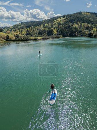 Two young girls enjoying stand-up paddleboarding on a river surrounded by lush nature on a bright summer day. Embracing outdoor adventure and activities in the natural beauty of their surroundings