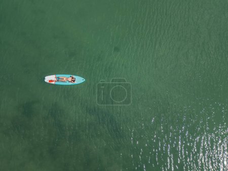 Photo for Cenital young brunette Latina resting and sunbathing while lying on her surfboard in the middle of the sea on sunny summer day. Enjoying the relaxed atmosphere and the warm waters of the ocean - Royalty Free Image