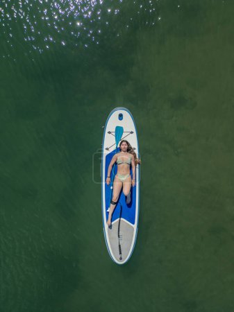 Photo for Cenital vertical young brunette Caucasian woman resting and sunbathing while lying on her surfboard in the middle of the sea on a hot summer day. Enjoying the warmth of the sun and the tranquil waters - Royalty Free Image