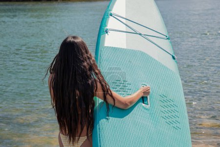 hispanic girl in a bikini, seen from behind, carrying her paddle surf board towards the water of the lake on a summer vacation day. Enjoying outdoor water activities and leisure time in the sun
