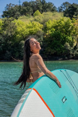 smiling Latin woman in a bikini holding her paddleboard while the sun shines on her face on a summer day. Enjoying outdoor water activities and soaking up the sun
